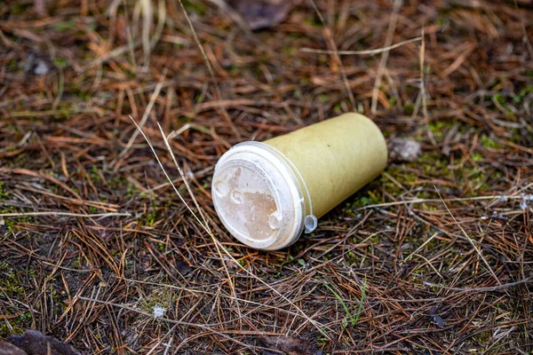 A plastic cup is on the ground in a field. The cup is empty and has a lid on it. The scene is desolate and abandoned, with no one around to clean up the trash