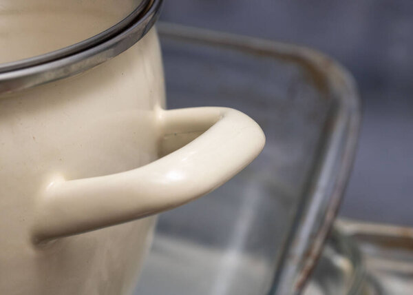 A white pot with a silver handle sits on a table. The pot is on a glass surface, and the handle is visible. Concept of warmth and comfort, as the pot is likely used for cooking or serving food