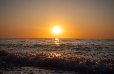 Güneş okyanusun üzerinde batıyor, suyun üzerine sıcak bir parıltı saçıyor. Gökyüzü turuncu ve pembe renklerin bir karışımıdır. Huzurlu ve huzurlu bir atmosfer yaratır. Dalgalar yavaşça kıyıya vurur