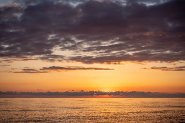 Gökyüzü bulutlu ve güneş batıyor. Okyanus sakin ve su koyu mavi bir renktir.