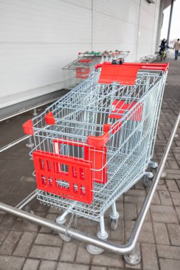 Bir mağazanın önündeki kaldırımda bir alışveriş arabası oturuyor. Araba boş ve kırmızı saplı.