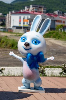 Ahşap bir platformda duran mavi ve pembe bir tavşan heykeli. Tavşan gülümsüyor ve boynunda mavi bir fiyonk var. Heykel, çimenler ve ağaçlarla çevrili bir parkta bulunuyor.