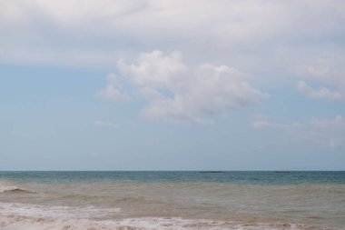 Arka planda büyük bir su kütlesi olan bulutlu bir gökyüzü. Gökyüzü çoğunlukla mavi ve birkaç bulut etrafa saçılmış. Su sakin ve kumsal boş.