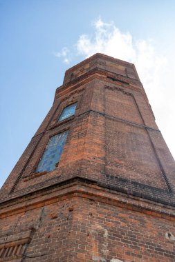 Üzerinde saat olan uzun bir tuğla kule. Kule tuğla bir duvarla çevrilidir. Gökyüzü mavi ve berrak