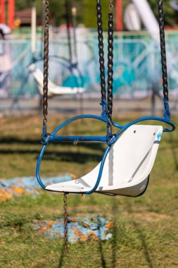 Mavi ve beyaz bir salıncak zincirden sarkıyor. Salıncak boş ve koltuk kayıp. Salıncak bir parkta ve arka planda başka salıncaklar var.