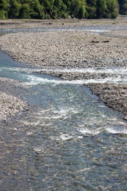 Kayalık bir kıyı şeridi olan bir nehir. Su berrak ve sakin. Kayalar nehir boyunca dağılıp doğal bir bariyer oluşturuyor. Huzurlu ve huzurlu bir sahne.