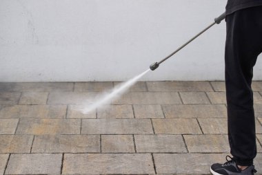 Bir kişi tuğla zemini temizlemek için basınç makinası kullanıyor. Basınçlı çamaşır makinesi yere su püskürtüyor ve o kişi de yakınlarda duruyor. Temizlik ve bakım kavramı