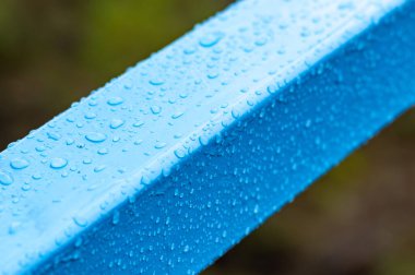 Üzerinde su damlacıkları olan mavi metal bir direk. Damlacıklar küçük ve dağınık, hareket ve enerji hissi yaratıyorlar. Kutbun mavi rengi yeşil arkaplanla çelişiyor