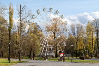 Ortasında büyük beyaz dönme dolap olan bir park. Park boş ve gökyüzü bulutlu.