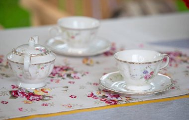 Üzerinde çay seti olan bir masa. Masa örtüsünün çiçek deseni var. Çay takımında çaydanlık, fincan ve çay tabağı var.