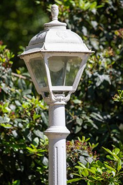 İçinde ampul olan beyaz bir lamba direği. Lamba direği yeşil çalılarla çevrili. Resmin huzurlu ve huzurlu bir havası var.