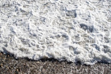 Büyük bir dalga kayalık bir sahile çarpıyor. Su beyaz ve köpüklü ve kayalar sahnenin her tarafına saçılmış. Manzara güçlü ve dinamiktir.