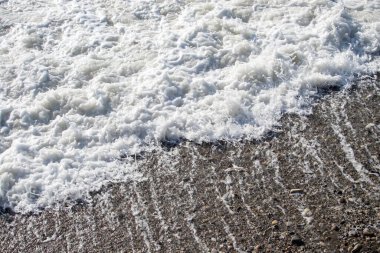 Büyük bir dalga kayalık bir sahile çarpıyor. Su beyaz ve köpüklü ve kayalar sahnenin her tarafına saçılmış. Manzara güçlü ve dinamiktir.