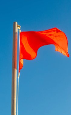 Açık mavi gökyüzünde rüzgarda kırmızı bir bayrak dalgalanıyor. Bayrak direğin üzerinde ve görüntüde görünen tek şey o.
