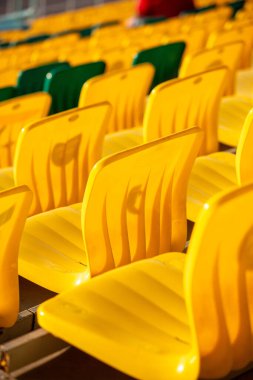 Stadyumda bir sıra sarı sandalye boş. Sandalyeler sıra sıra diziliyor, bazı sandalyeler kameraya bakıyor ve diğerleri diğer tarafa bakıyor. Sandalyelerin hepsi aynı renk ve şekildedir.