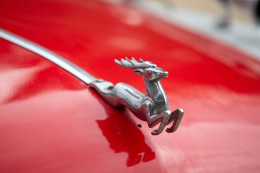 Kırmızı bir arabada gümüş bir geyik var. Geyik kameraya bakıyor. Araba bir caddeye park edilmiş.