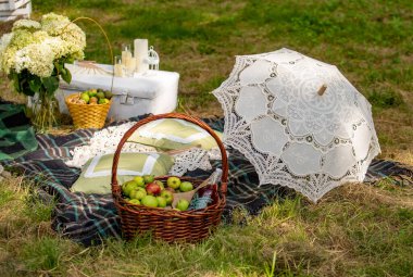 A picnic basket full of apples and a white lace umbrella are on a blanket in a grassy field