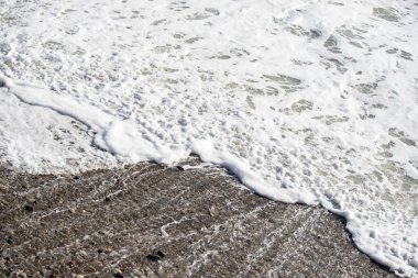 Bir dalga kayalık bir sahile çarpıyor. Su beyaz ve köpüklü ve kayalar sahnenin her tarafına saçılmış. Sahne sakin ve huzurlu, dalgalar yavaşça kıyıya vururken
