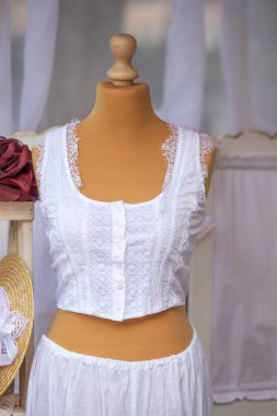 Bir kadının beyaz atlet bluzu ve beyaz pantolonu bir mankenin üzerinde sergilenir. Kolsuz tişörtte dantel temizleme ve önünde bir düğme var. Sahne zarif ve kadınsı.