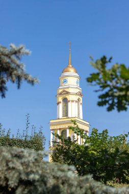 Mavi gökyüzünün önünde altın kapağı olan uzun sarı bir kule duruyor. Kule ağaçlarla çevrili ve yapraklar yeşil. Sahne huzurlu ve huzurlu, kule dik duruyor.