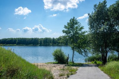 Önünde bir patika olan güzel bir göl. Gökyüzü bazı bulutlarla mavidir. Ağaçlar yeşil ve su sakin.