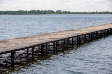 Tahta bir köprü bir su kütlesini kapsar. Su sakin ve gökyüzü bulutlu. Köprü eski ve yıpranmış, ama hala sağlam.