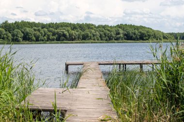 Bir rıhtım göl kenarında, deniz manzaralı. Rıhtım ahşap ve ona giden bir köprüsü var. Su sakin ve gökyüzü bulutlu.