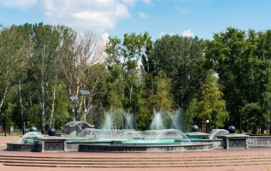 Arka planda ağaçlar olan bir parkta çeşme. Çeşme merdivenlerle çevrili ve yeşil bir rengi var.