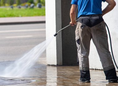 Bir adam kaldırımı hortumla yıkıyor. Kaldırımlar ıslak ve adam onu temizlemek için basınç makinası kullanıyor.