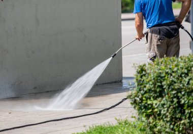 Bir adam bir hortumla duvarı yıkıyor. Hortum duvara su püskürtüyor. Adam mavi gömlek ve gri şort giyiyor.