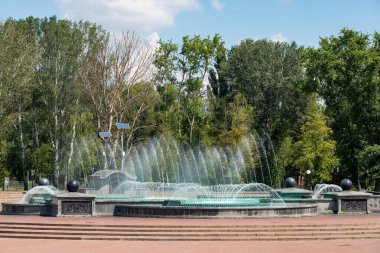 Havaya fışkıran suyla dolu bir çeşme. Çeşme ağaçlarla çevrili ve taştan bir tabanı var.
