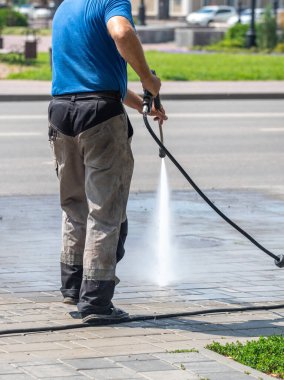 Bir adam kaldırımı hortumla yıkıyor. Kaldırımlar ıslak ve adam onu temizlemek için basınç makinası kullanıyor.