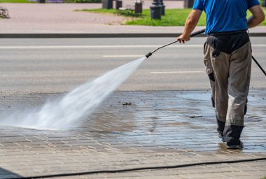 Bir adam kaldırımı hortumla yıkıyor. Kaldırımlar ıslak ve adam çok fazla su kullanıyor.