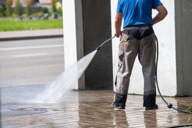 Bir adam kaldırımı hortumla yıkıyor. Kaldırımlar ıslak ve kirli. Adam mavi gömlek ve gri pantolon giyiyor.