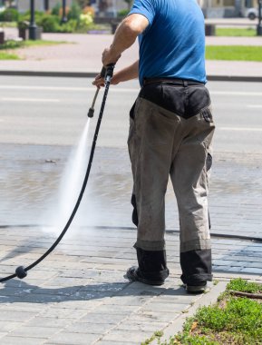 Bir adam kaldırımı hortumla yıkıyor. Kaldırımlar kirli ve adam onu temizlemek için çamaşır makinesi kullanıyor.