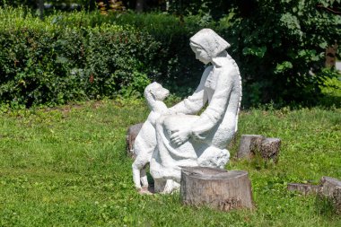 Kütüğün üzerinde oturan bir kadın heykeli ve bir köpek. Heykel beyaz ve köpek küçüktür. Huzurlu ve huzurlu bir sahne.