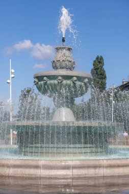 İçinden su fışkıran büyük bir çeşme. Su temiz ve çeşme bir taş tabanla çevrili. Çeşme bir parkta ve ağaçlarla çevrili.