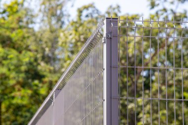 A metal fence with a wire mesh on top. The fence is surrounded by trees and bushes. The fence is tall and has a sharp edge
