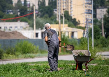 Bahçede el arabası olan bir adam çalışıyor. Adam gri bir gömlek ve gri pantolon giyiyor.