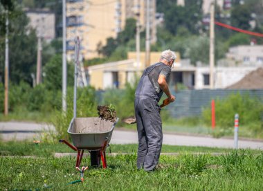 Bahçede el arabası olan bir adam çalışıyor. Adam gri bir gömlek ve gri pantolon giyiyor.