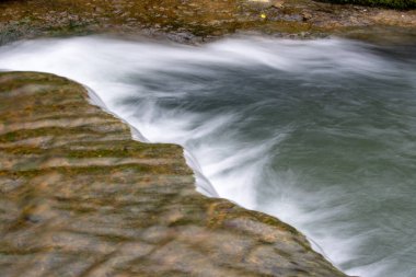 Kayalık bir çıkıntıdan akan bir su akıntısı. Su berrak ve sakin, ve kayalar sahnenin her tarafına dağılmış. Sahne huzurlu ve sakin, su kayaların üzerinden yavaşça akıyor.
