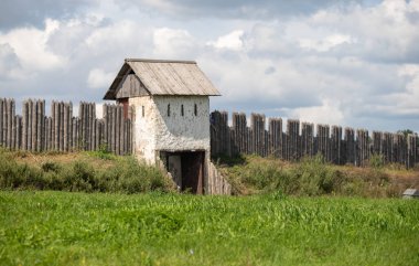 A small house with a stone roof sits in a field. The house is surrounded by a wooden fence. The sky is cloudy and the grass is green