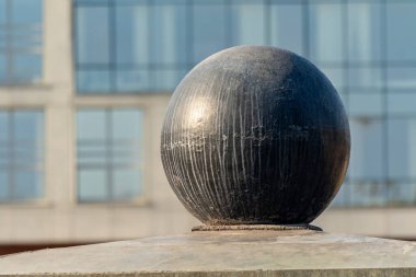 Büyük yuvarlak metal bir top bir çıkıntının üzerinde duruyor. Top metal bir çerçeve ile çevrilidir ve beton bir yüzey üzerinde oturmaktadır. Sahne, geniş pencereli bir binanın arka planına göre ayarlanmış.