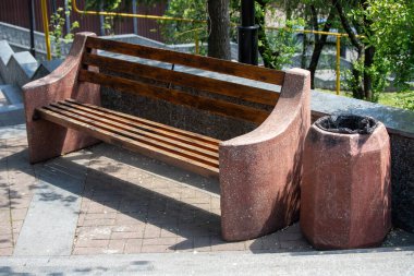 Bir bankta ve bir çöp tenekesi tuğla kaldırımda oturuyor. Banka ahşaptan yapılmıştır ve üzerinde sarı bir şerit vardır. Çöp kutusu betondan yapılmış ve siyah bir kapağı var. Sahne huzurlu ve sessiz.
