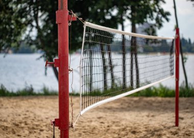 A volleyball net is set up on a sandy beach near a body of water. The net is red and white, and it is leaning against a pole. The scene is peaceful and relaxing, with the water