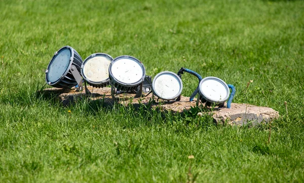 A row of lights are on a grassy surface. The lights are of different sizes and are arranged in a line. The grass is green and the sky is blue