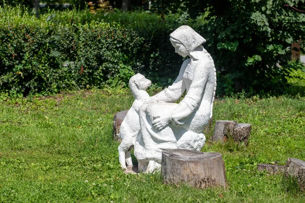 Kütüğün üzerinde oturan bir kadın heykeli ve bir köpek. Heykel beyaz ve köpek küçüktür. Huzurlu ve huzurlu bir sahne.