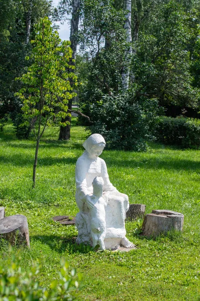 Parktaki bankta oturan bir kadın ve bir çocuk heykeli. Heykel beyaz ve park yeşil.