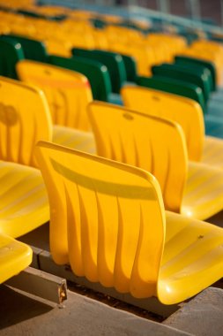 A row of yellow chairs are empty in a stadium. The chairs are arranged in a row, with some of them facing the camera. The chairs are all the same color, which gives the scene a uniform