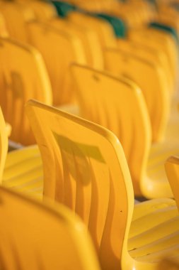 Bir sıra sarı sandalye boş. Sandalyeler sıraya dizilmiş, bazıları kameraya bakıyor. Sandalyelerin hepsi aynı renk ve boyutta ve hepsi boş.